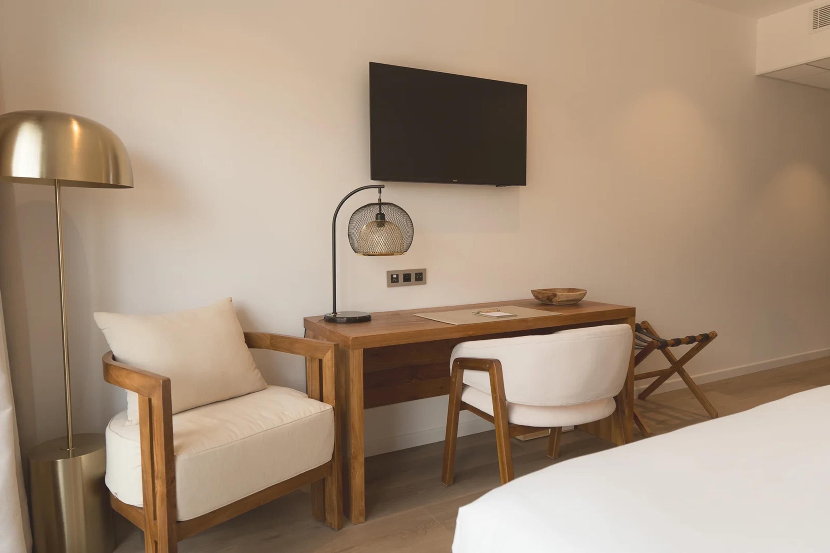 A modern hotel room with a wooden desk, a chair, a bedside table, a lamp, and a television mounted on the wall.