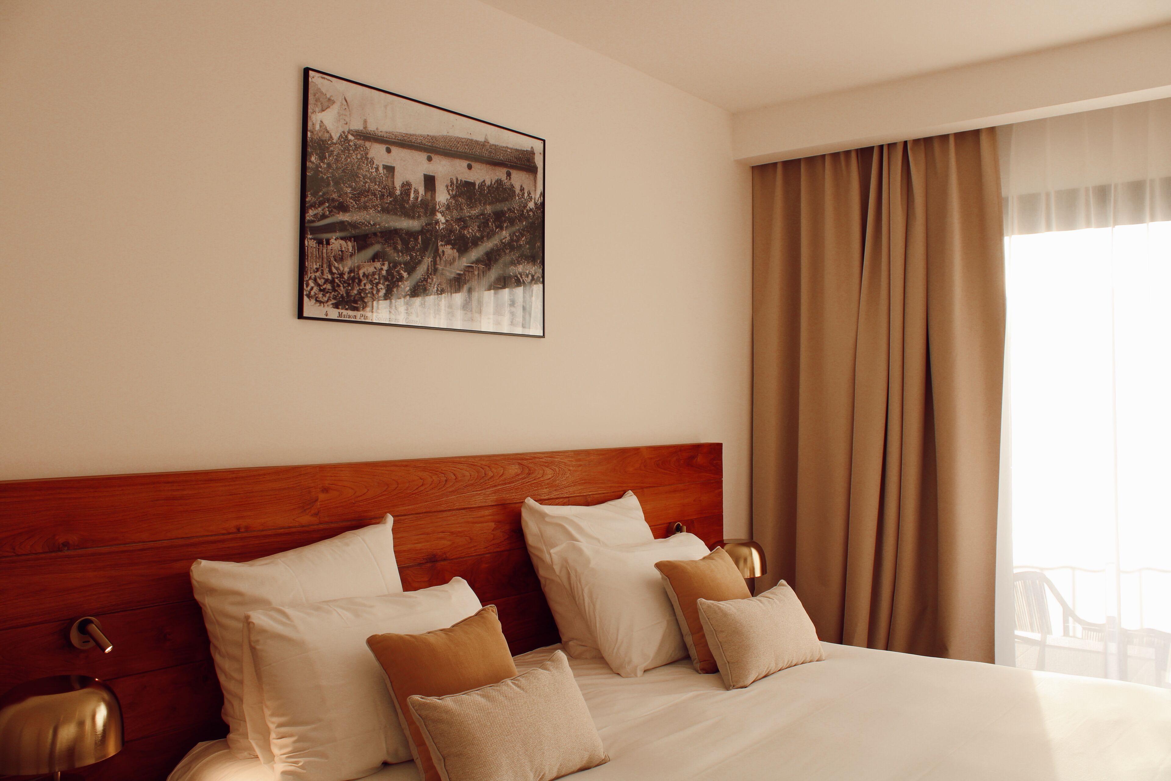 Hotelkamer met een houten hoofdbord, een groot bed met witte en beige kussens, een lijst met een afbeelding aan de muur, en een raam met gordijnen.