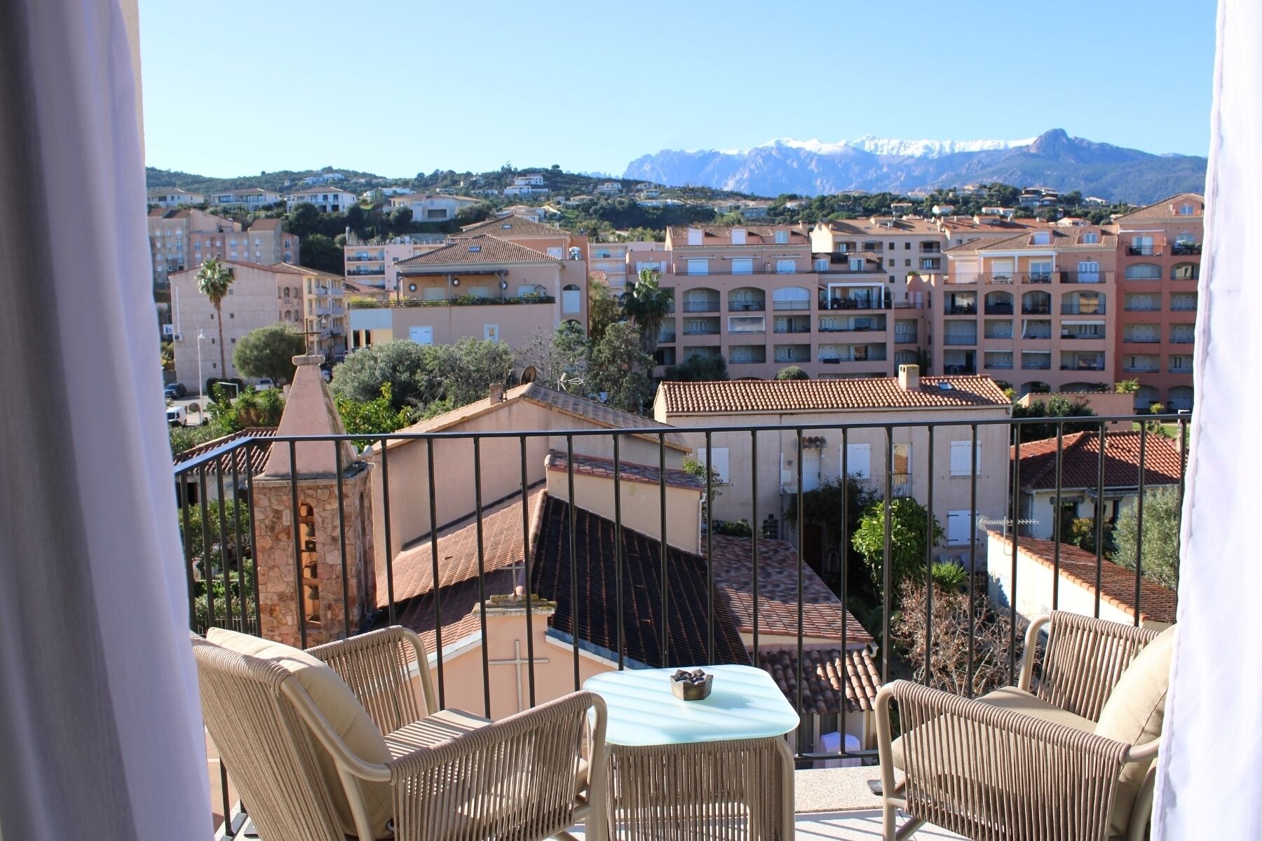 A balcony with wicker furniture and a table, overlooking a residential area with buildings and mountains in the background.