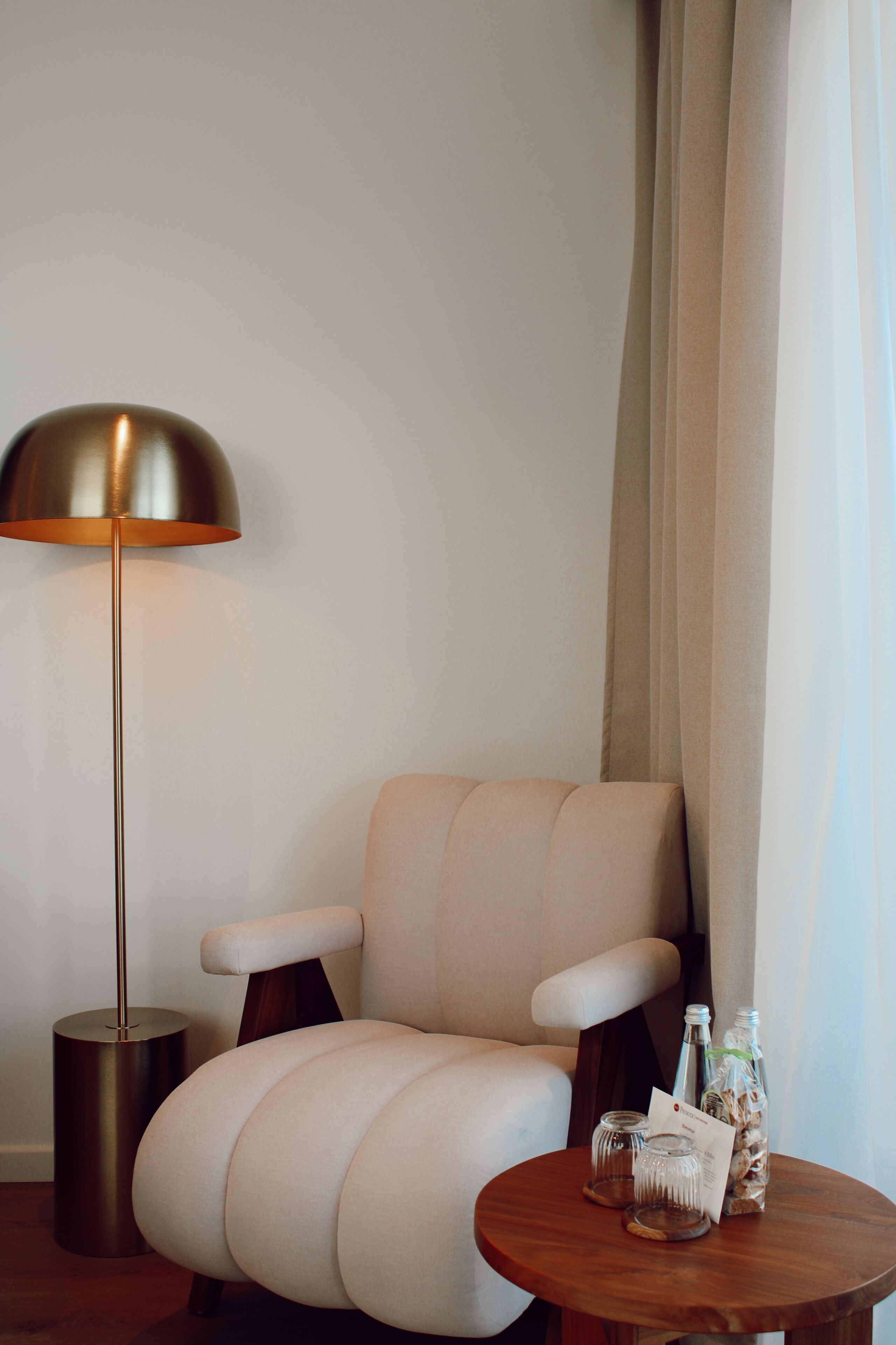 A modern armchair with a wooden base, a round wooden side table with a bottle and a glass, and a floor lamp.