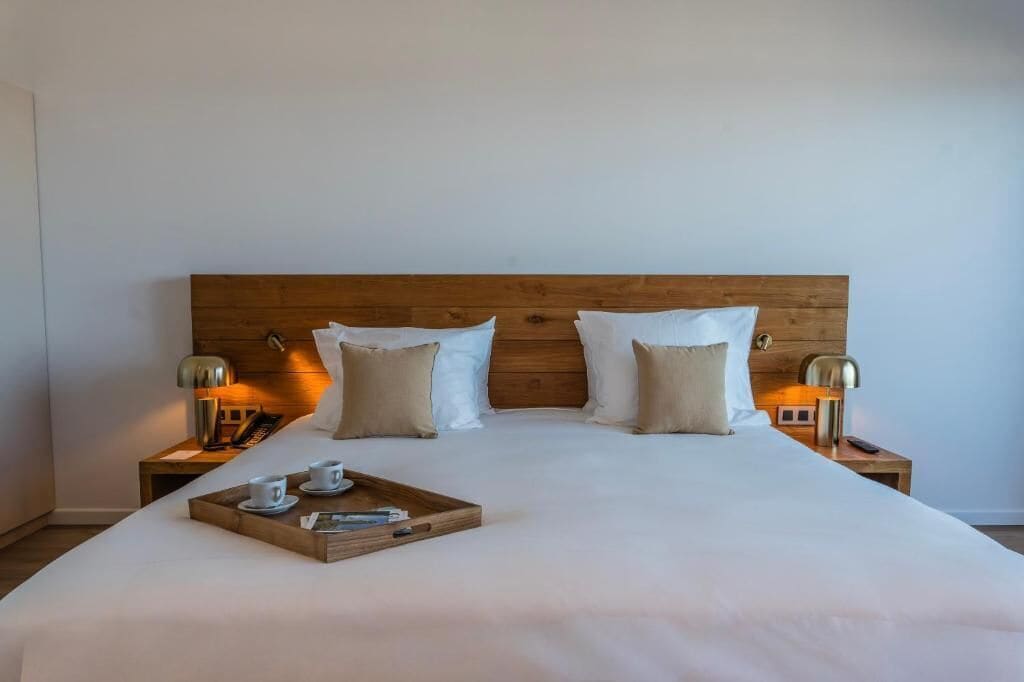 Una cama bien tendida con cabecera de madera, dos lámparas de noche y una bandeja con tazas y un libro sobre la cama.