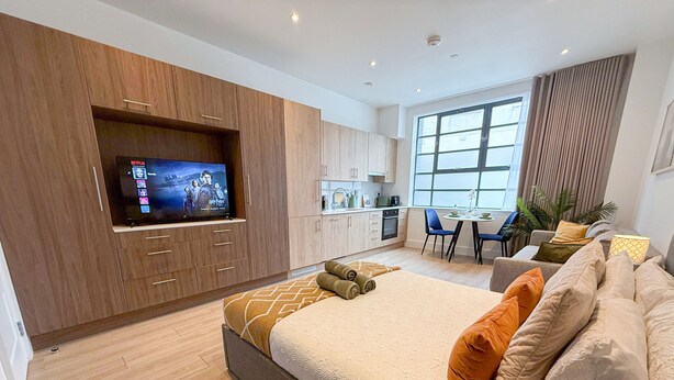 A modern bedroom with a large bed, a TV mounted on a wooden cabinet, a kitchenette, and a dining area with a large window.