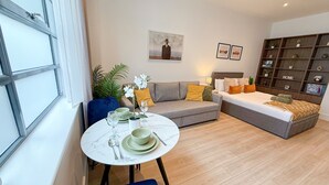 A modern living room with a dining area, a grey sofa, a blue chair, a round table with a white marble top, and a bookshelf with a 'HOME' sign.