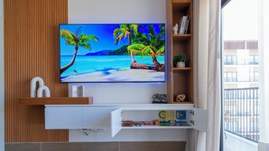 Ein modernes Wohnzimmer mit einem an einer Holzwand montierten Flachbildfernseher, einer weißen Unterhaltungseinheit darunter und einem großen Fenster mit Blick auf ein Gebäude.
