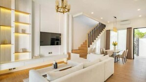 Una sala moderna con un sofá blanco grande, un televisor de pantalla plana montado en la pared, una escalera que sube a un nivel superior y un comedor con mesa y sillas.