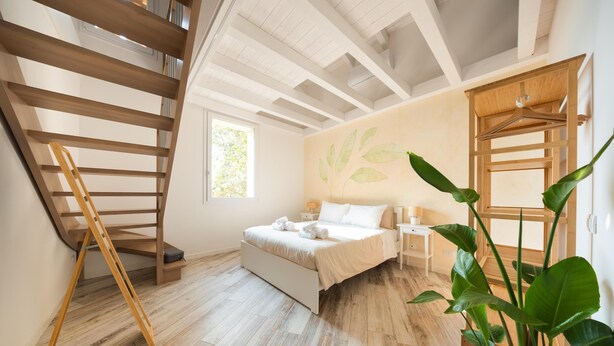 A bedroom with a wooden staircase, a bed with white linens, a nightstand, a bedside table, and a large plant. - Locanda L'orangerie (Crespellano)