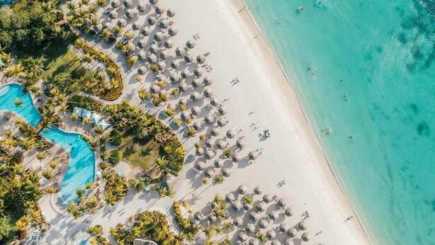 An aerial view of a beach resort with a swimming pool, palm trees, and beach umbrellas.