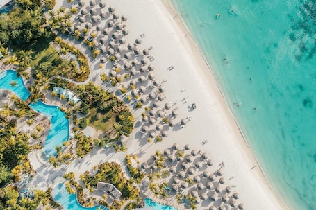 An aerial view of a beach resort with a swimming pool, palm trees, and beach umbrellas.