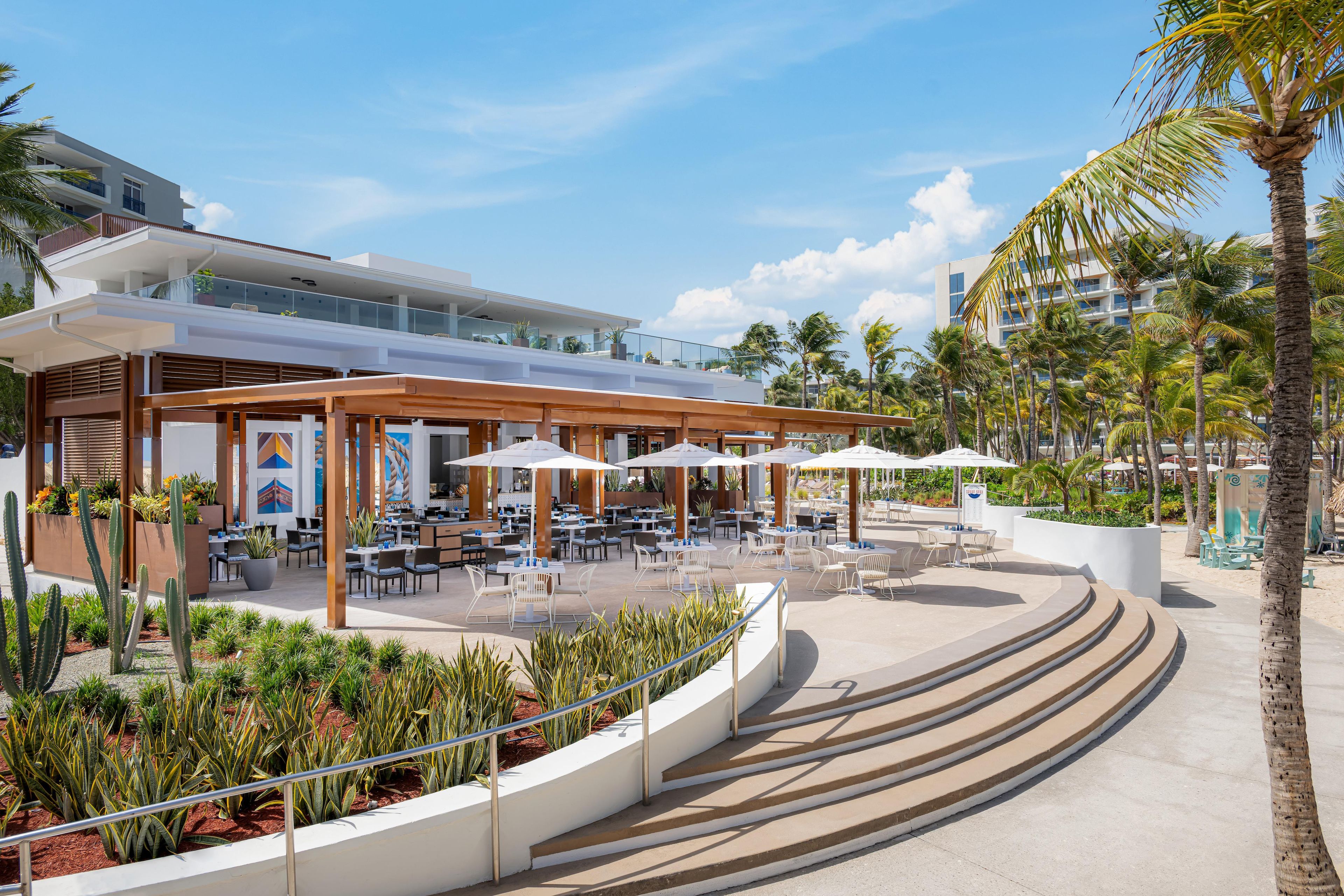 An outdoor dining area with tables and chairs, surrounded by palm trees and a modern building in the background.