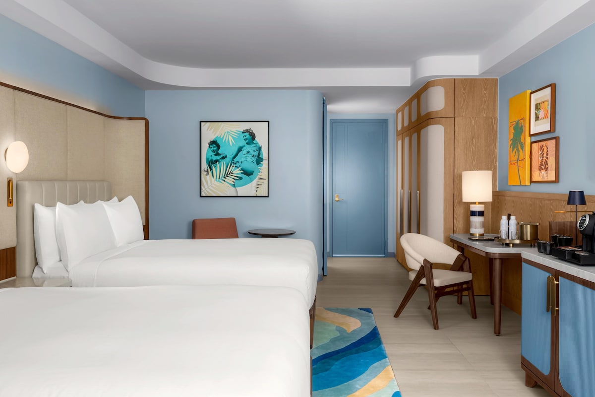 Quarto de hotel com cama, escrivaninha, cadeira, máquina de café e obras de arte nas paredes.