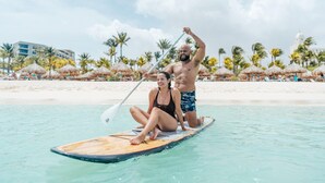 A couple on a paddleboard in a clear tropical beach setting with palm trees and thatched umbrellas.