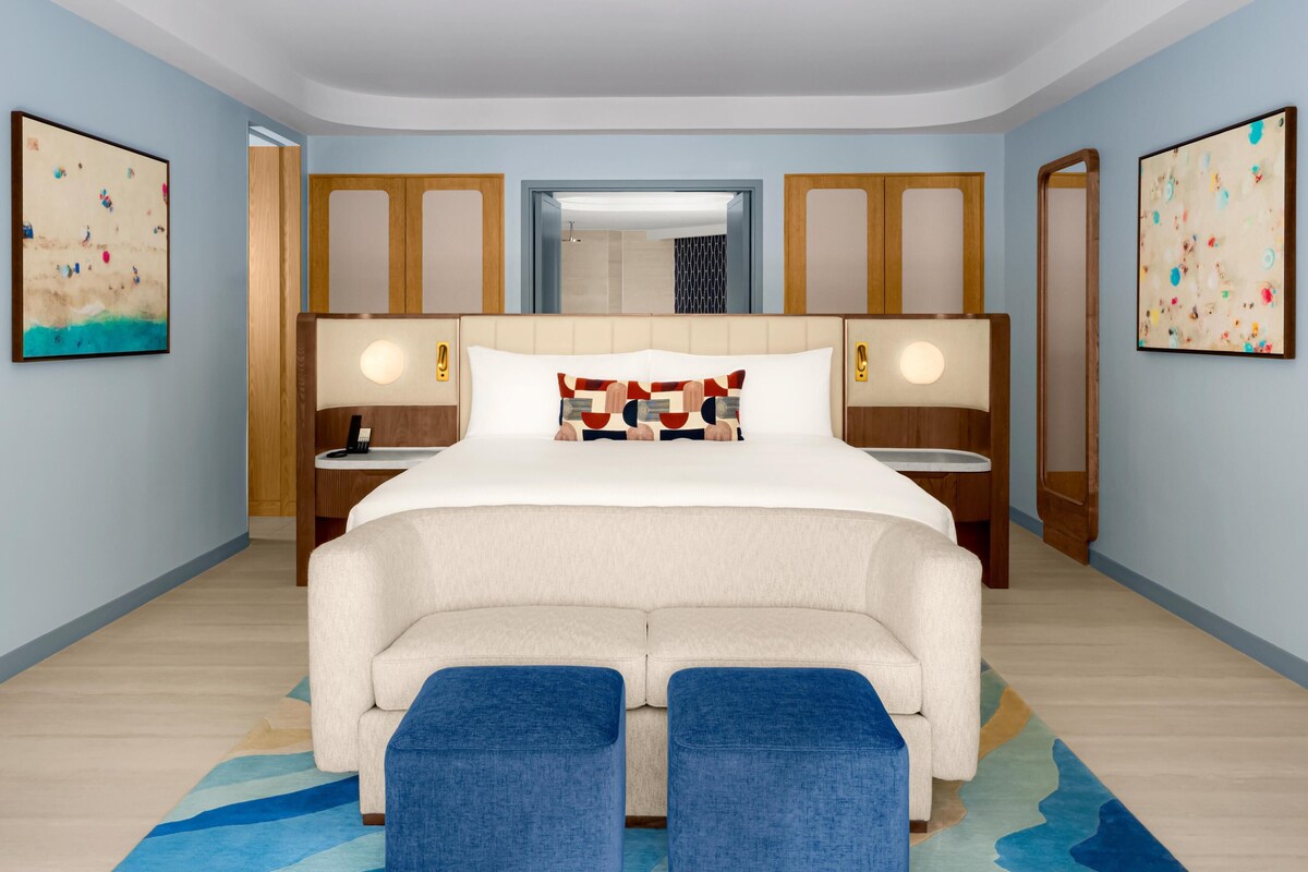 Une chambre d’hôtel avec un grand lit, deux tables de chevet, une commode, un miroir et des œuvres d’art accrochées aux murs.