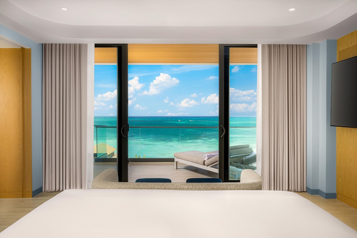 Quarto com uma cama grande, vista para o oceano e varanda com cadeira e mesa.