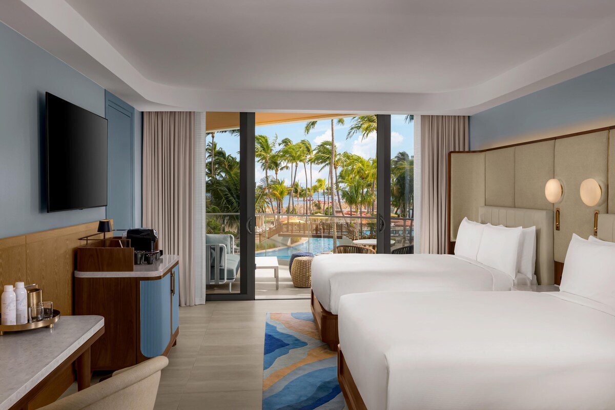 Quarto de hotel com cama, escrivaninha, televisão e vista para uma piscina e palmeiras.