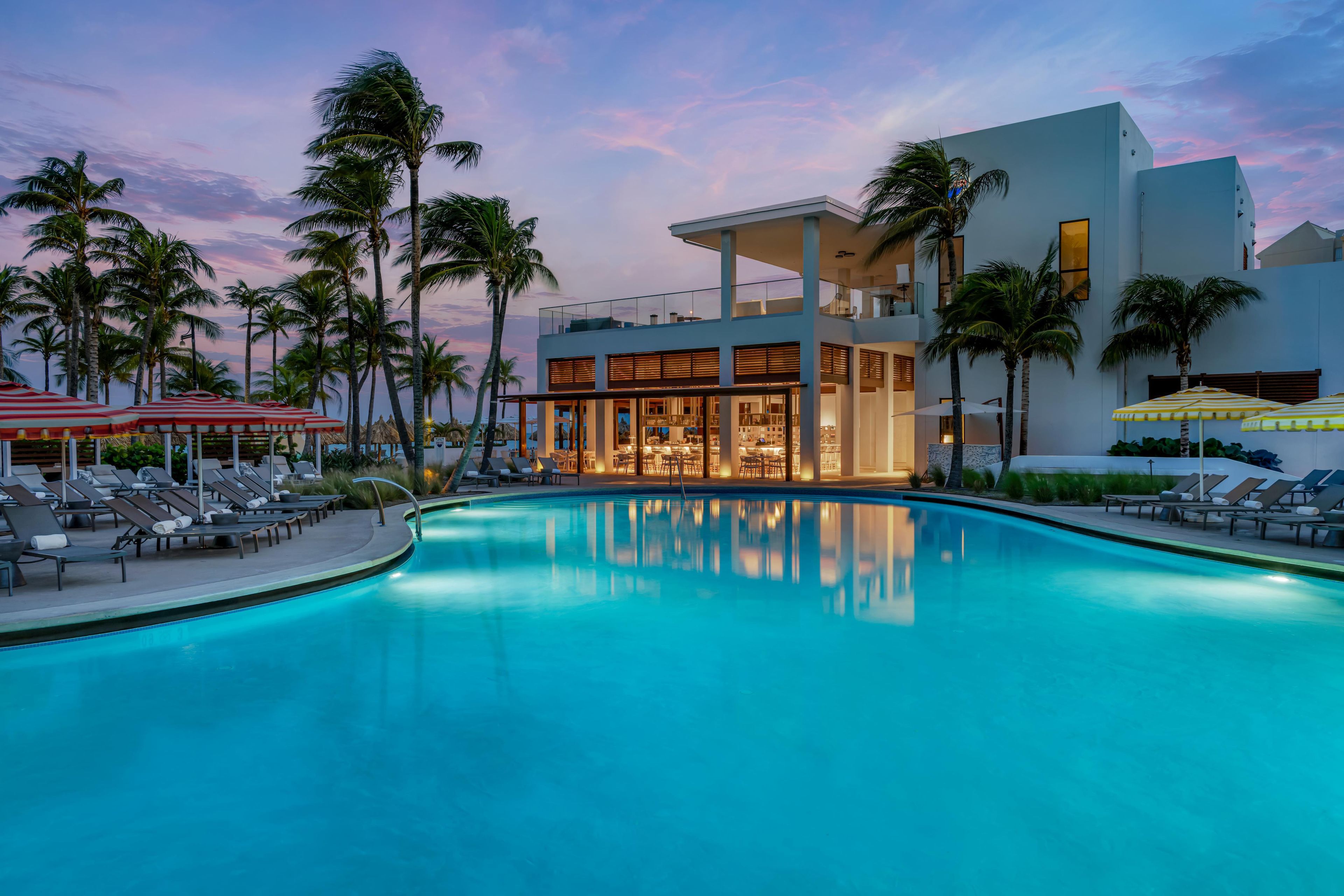 A modern hotel with a pool, lounge chairs, and palm trees.