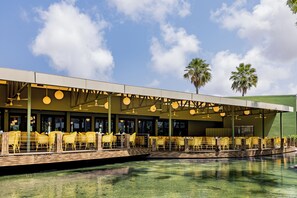 Un restaurante al aire libre con sillas y mesas amarillas, una piscina y palmeras.