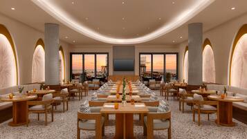 Un comedor amplio con varias mesas y sillas, ventanas grandes con vista al atardecer y una decoración interior moderna.