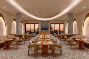 Un vaste espace dédié à la salle à manger, avec de nombreuses tables et chaises, de grandes fenêtres offrant une vue sur le coucher du soleil et un aménagement intérieur moderne.
