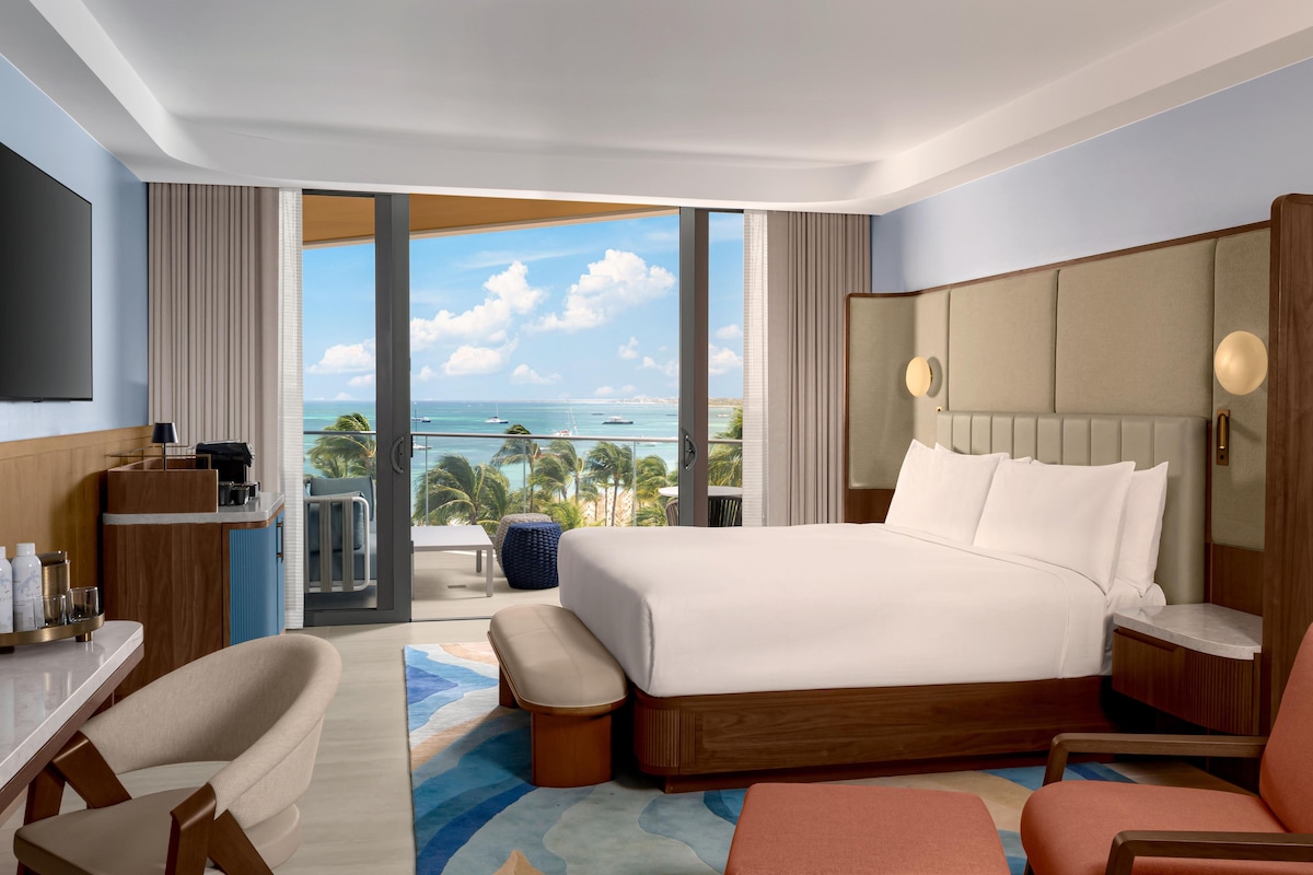 Quarto de hotel com uma cama grande, uma escrivaninha com televisão de tela plana, área de estar com vista para o mar e varanda com vista para a praia.