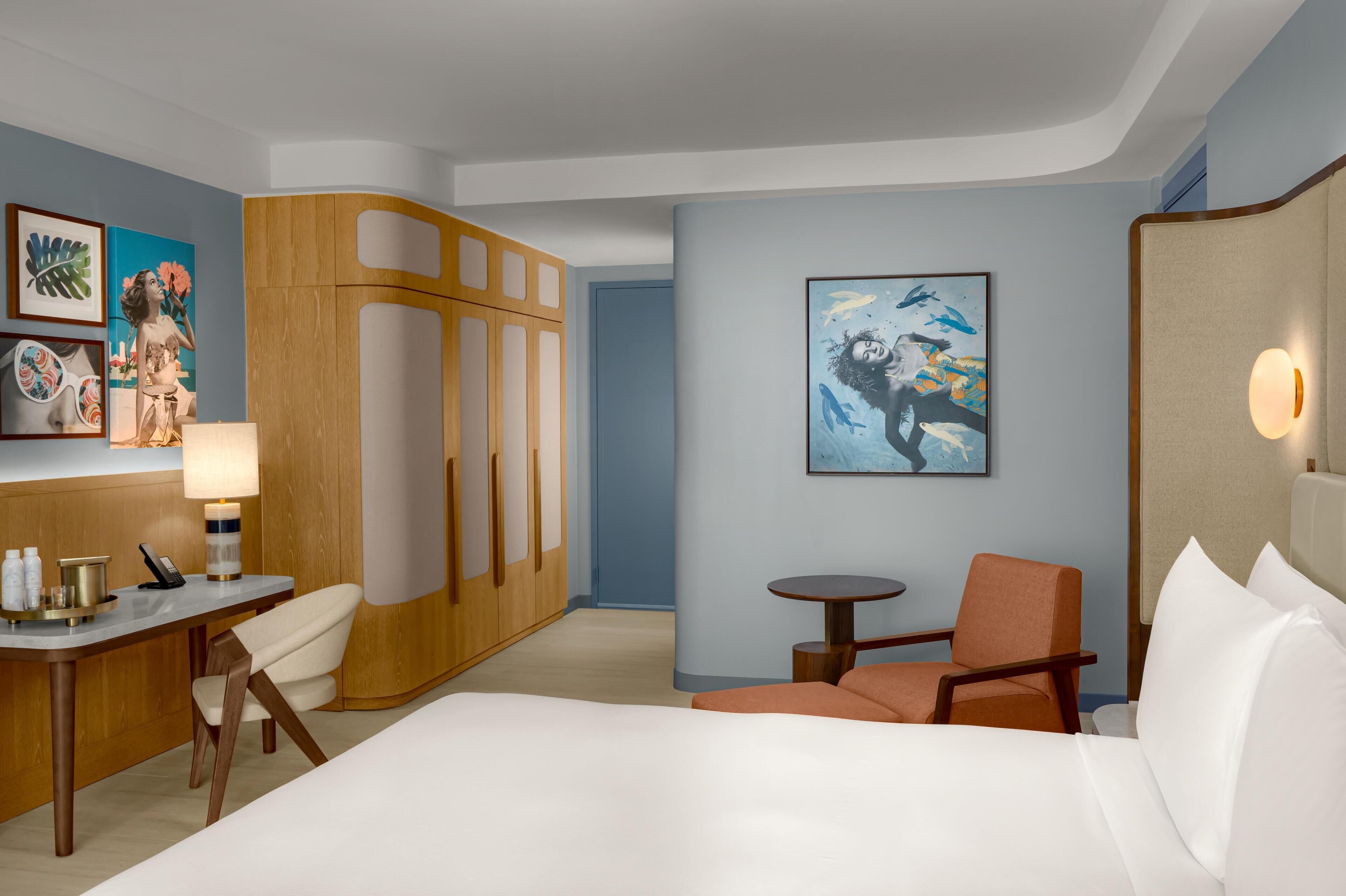 Habitación de hotel con cama, escritorio, silla, armario y obras de arte en las paredes.