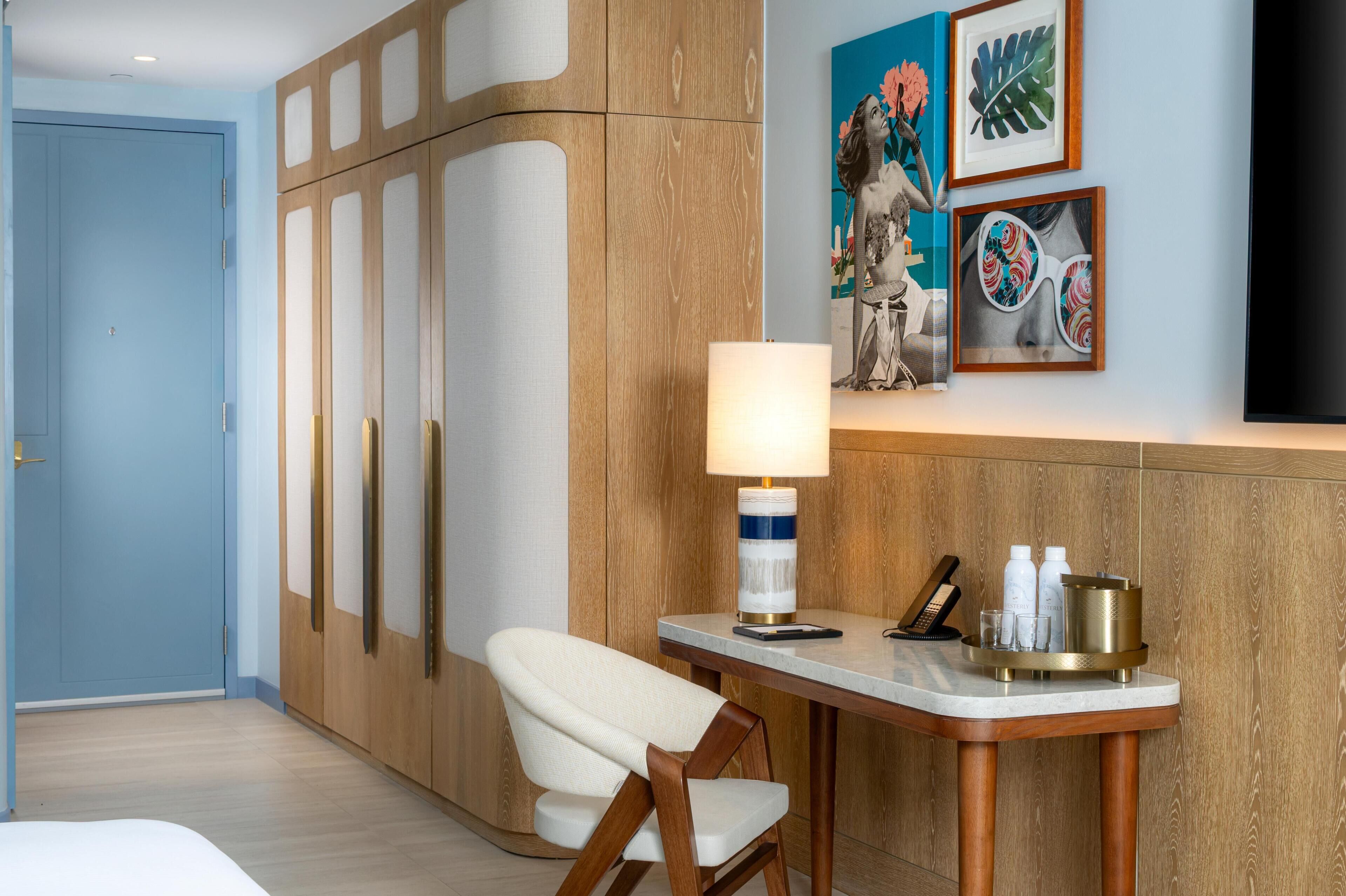 A hotel room with a bed, a desk with a lamp, a chair, a wardrobe, and artwork on the wall.