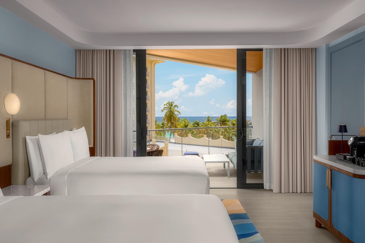 Quarto de hotel com uma cama grande, uma escrivaninha com cafeteira e uma varanda com vista para palmeiras e o oceano.
