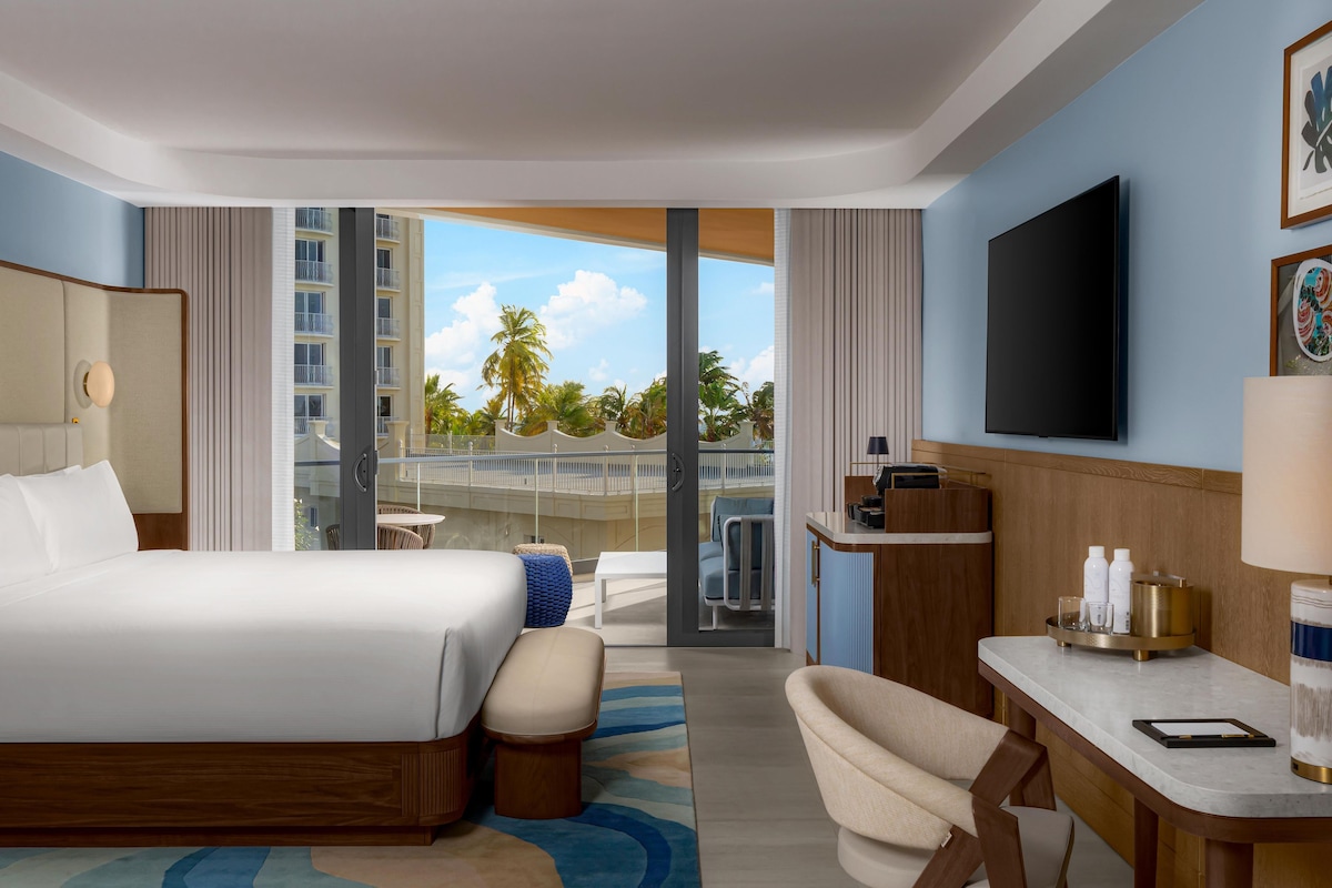 Quarto de hotel com cama, escrivaninha, cadeira, televisão e varanda com vista para palmeiras.
