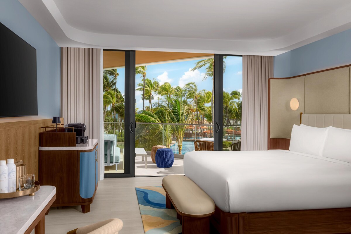 Une chambre d’hôtel avec un grand lit, un bureau doté d’une machine à café et une vue sur une piscine et des palmiers.