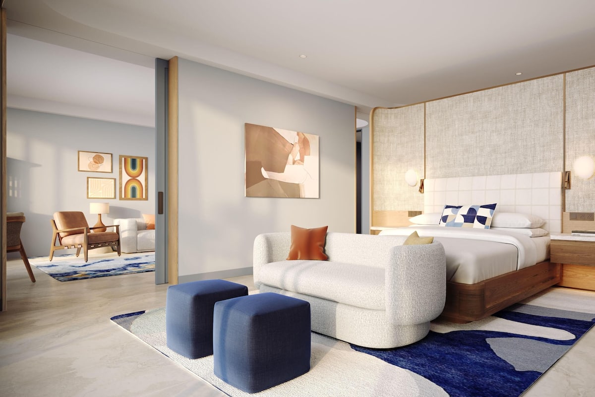 Ein modernes Schlafzimmer mit einem großen Bett, einer Sitzecke mit Poufs, einem Nachttisch und Kunstwerken an den Wänden.