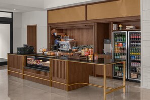 Una estación de café de autoservicio con diversas bebidas y aperitivos, un refrigerador lleno de refrescos y una barra con pasteles y aderezos.