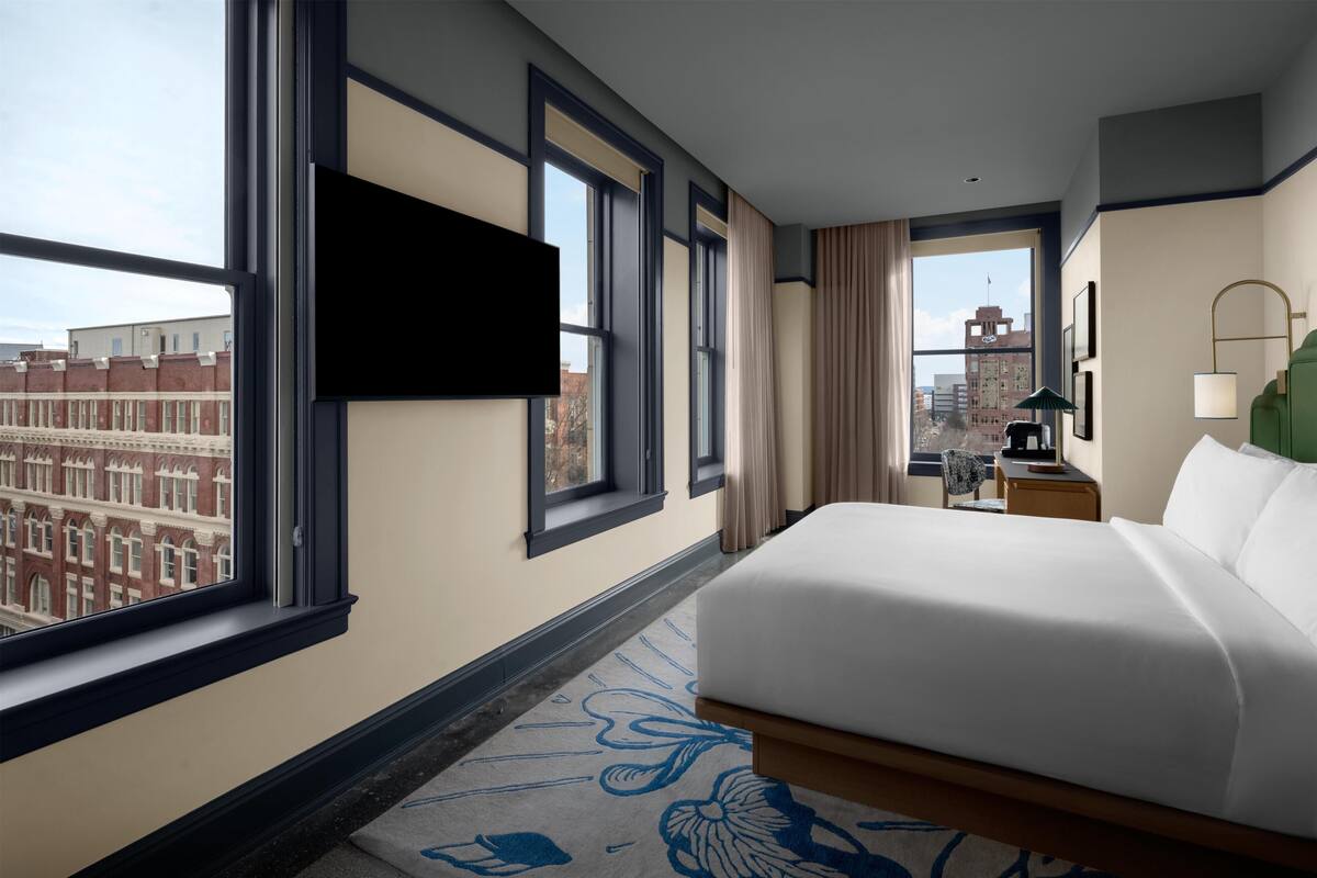 Une chambre d’hôtel dotée d’un grand lit, d’un téléviseur à écran plat fixé au mur, d’un bureau avec une lampe et offrant une vue sur la ville grâce à de grandes fenêtres.