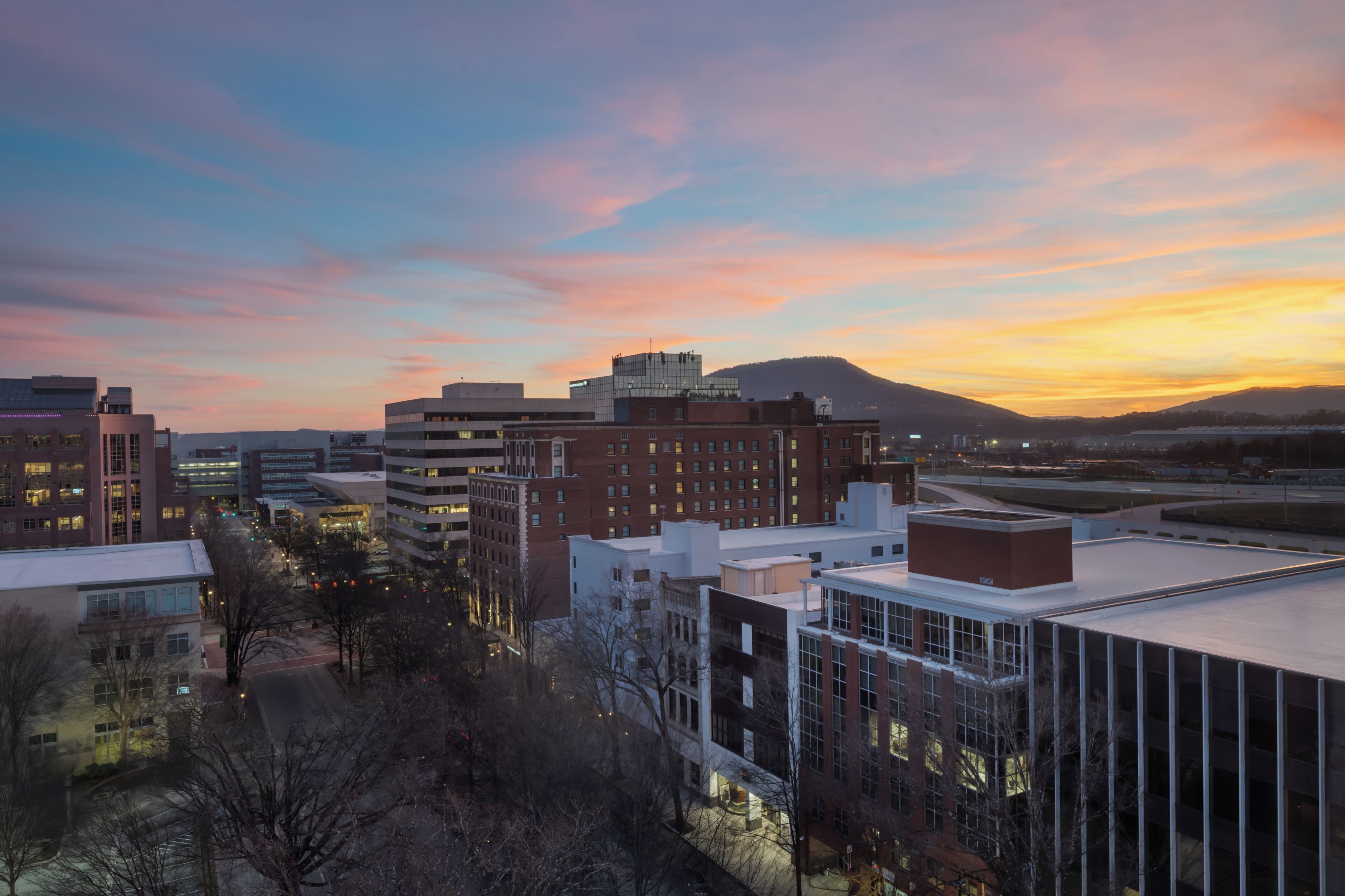 A cityscape at dusk with modern buildings, a clear sky, and distant hills.