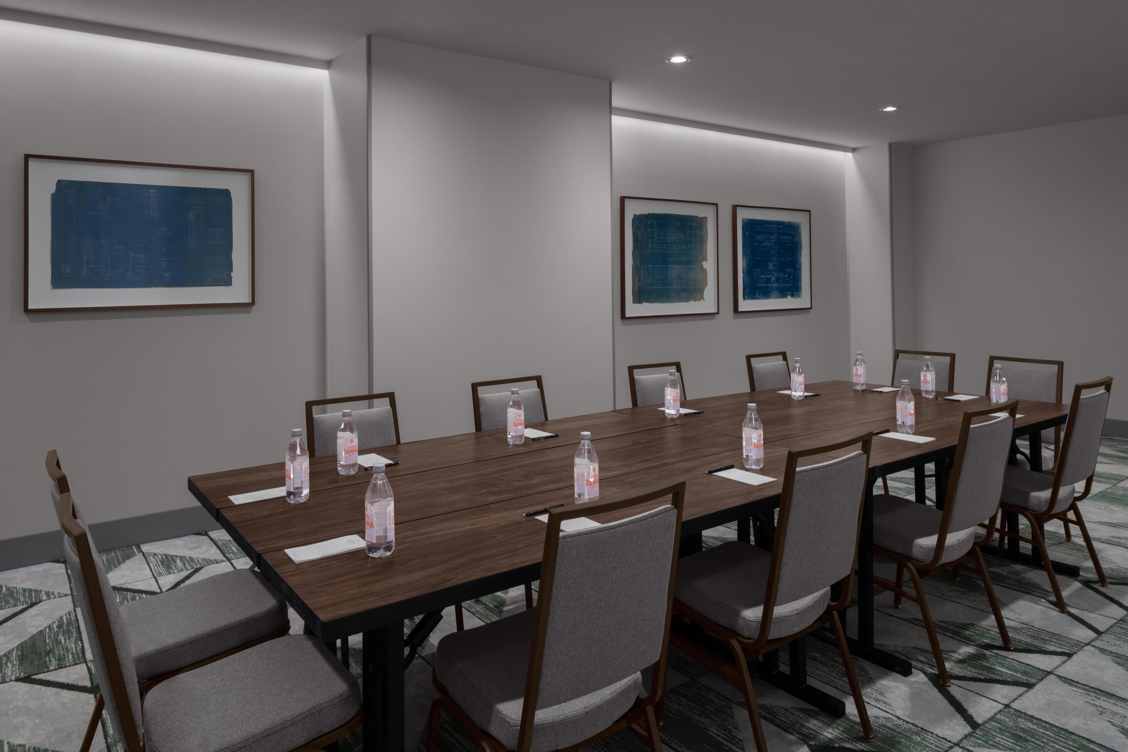 Uma sala de conferências com uma mesa comprida, cadeiras, obras de arte emolduradas nas paredes e garrafas de água sobre a mesa.
