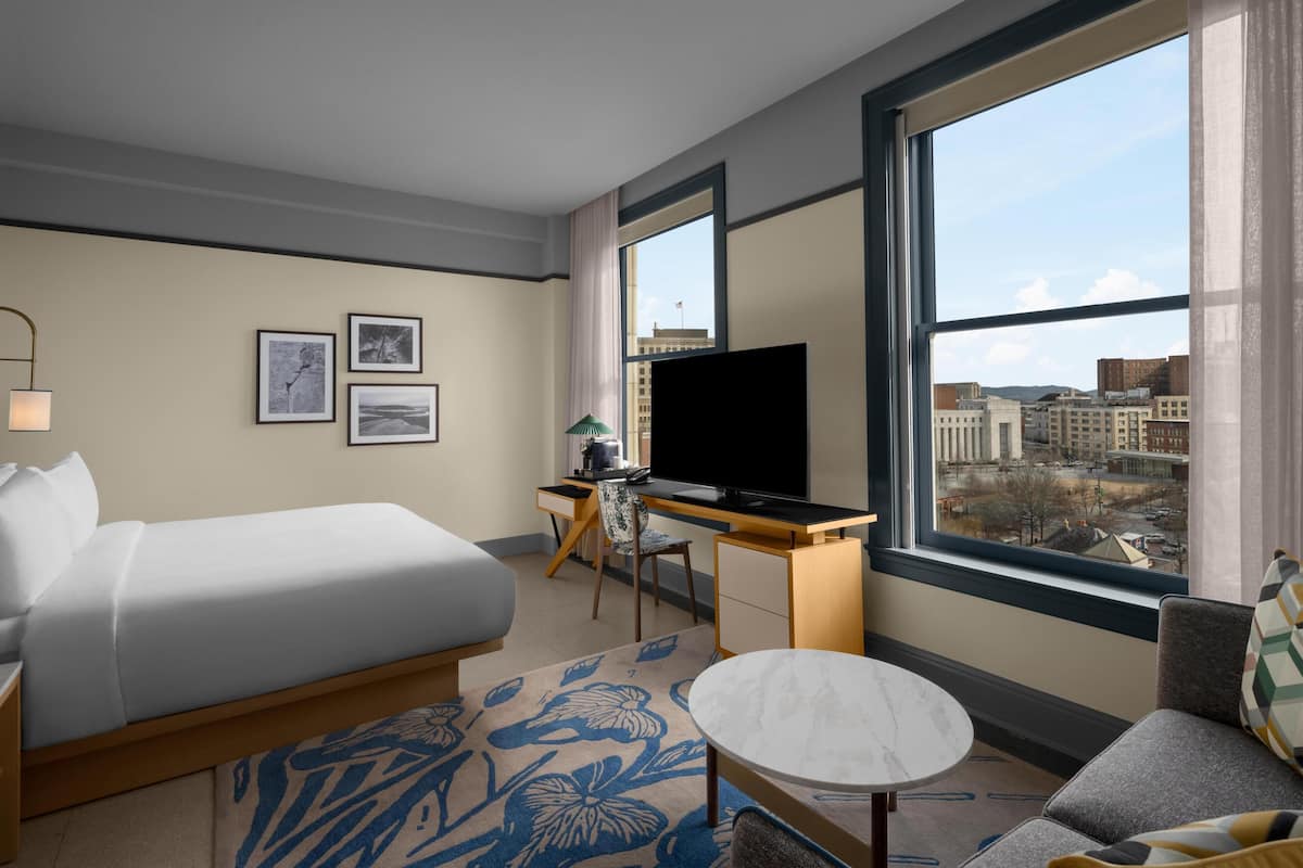 Une chambre d’hôtel avec un lit, un bureau avec une télévision, une chaise, une petite table et une grande fenêtre donnant sur la ville.