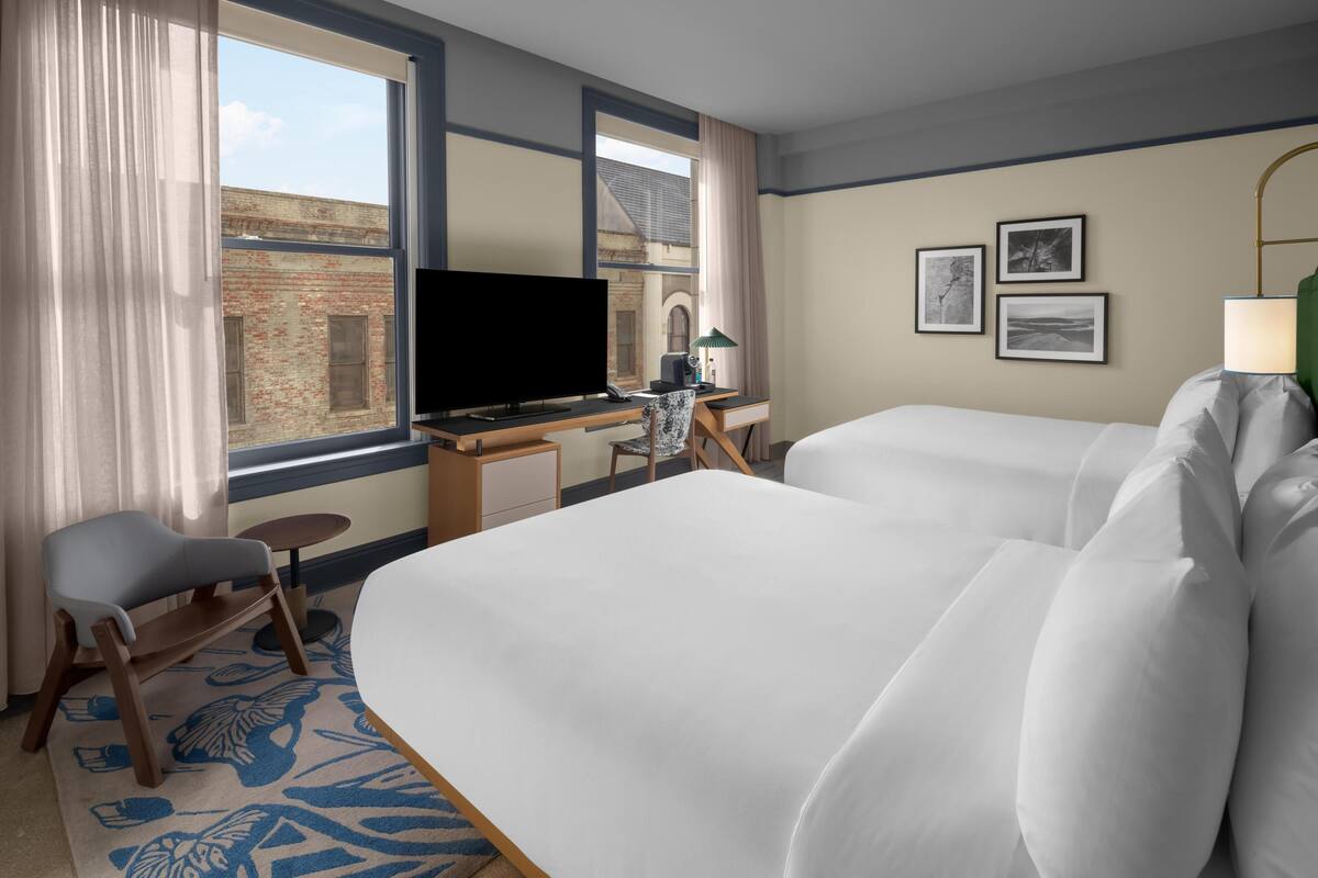Une chambre d’hôtel avec deux lits, un bureau avec un ordinateur, une chaise, une télévision et une fenêtre avec des rideaux.
