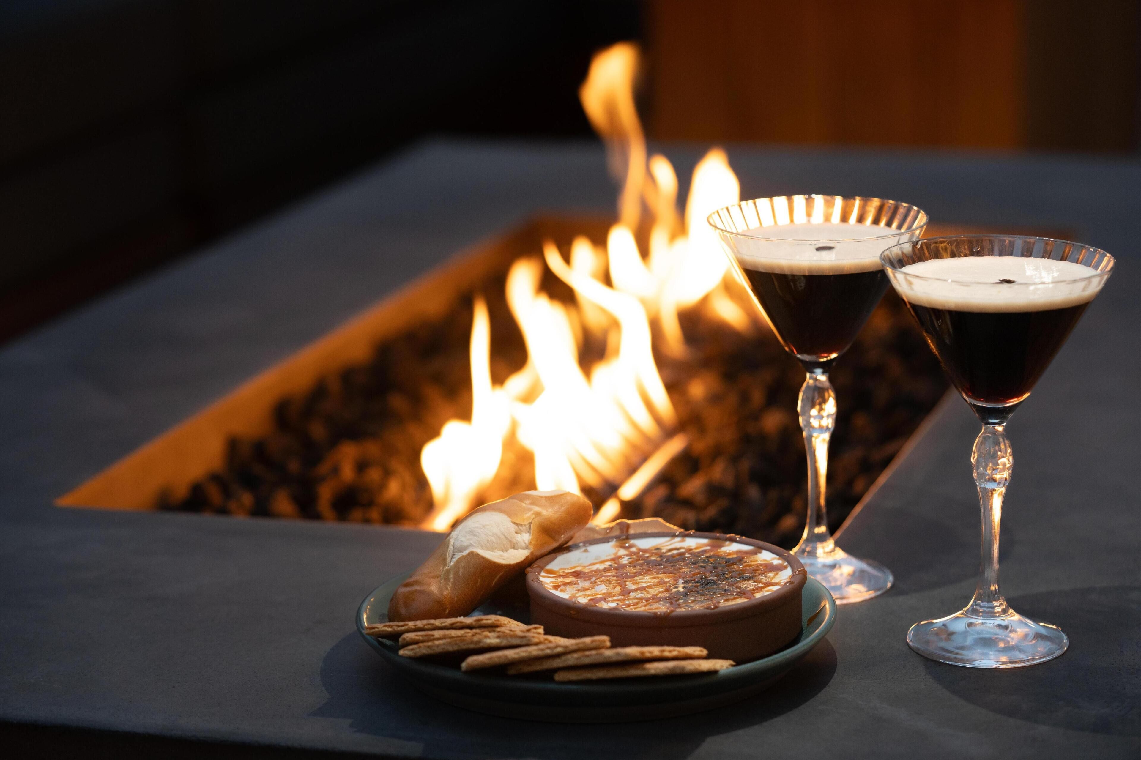 A table with a charcuterie board, two glasses of dark beverage, and a fire in the background.