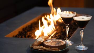 A table with a charcuterie board, two glasses of dark beverage, and a fire in the background.