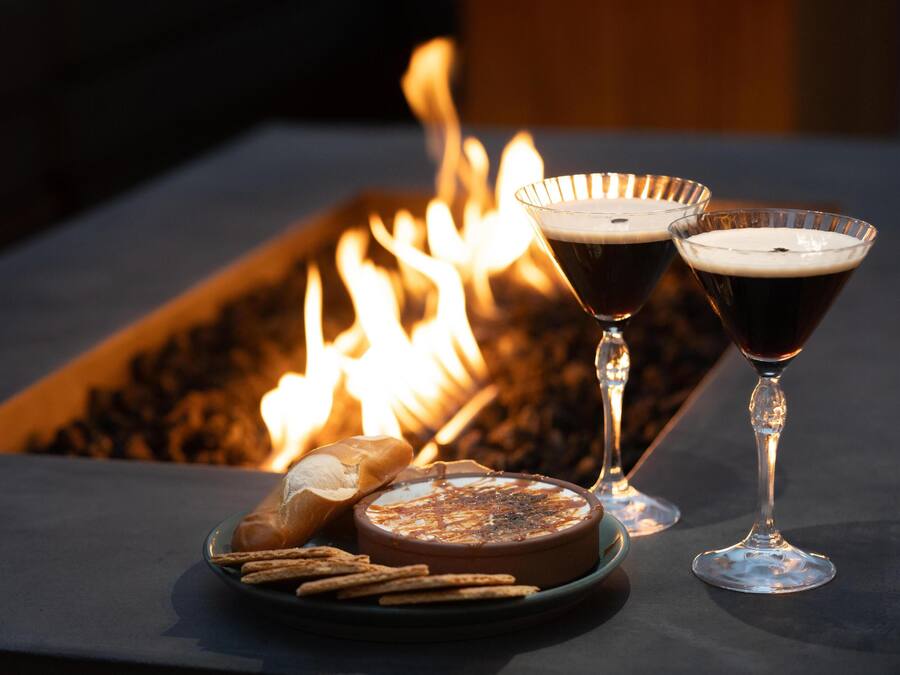 A table with a charcuterie board, two glasses of dark beverage, and a fire in the background.