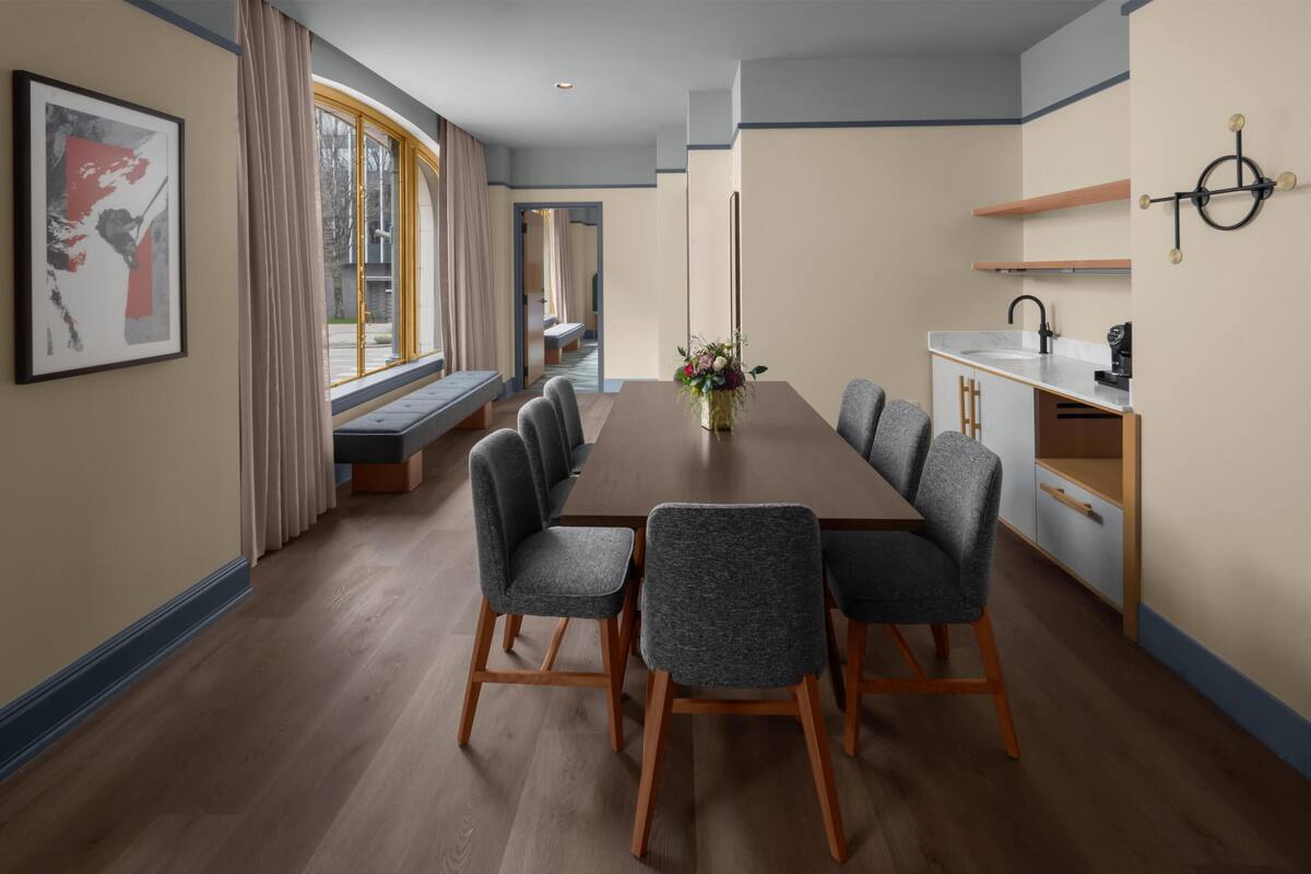 Un coin repas avec une longue table, six chaises, un vase de fleurs et une fenêtre avec des rideaux.