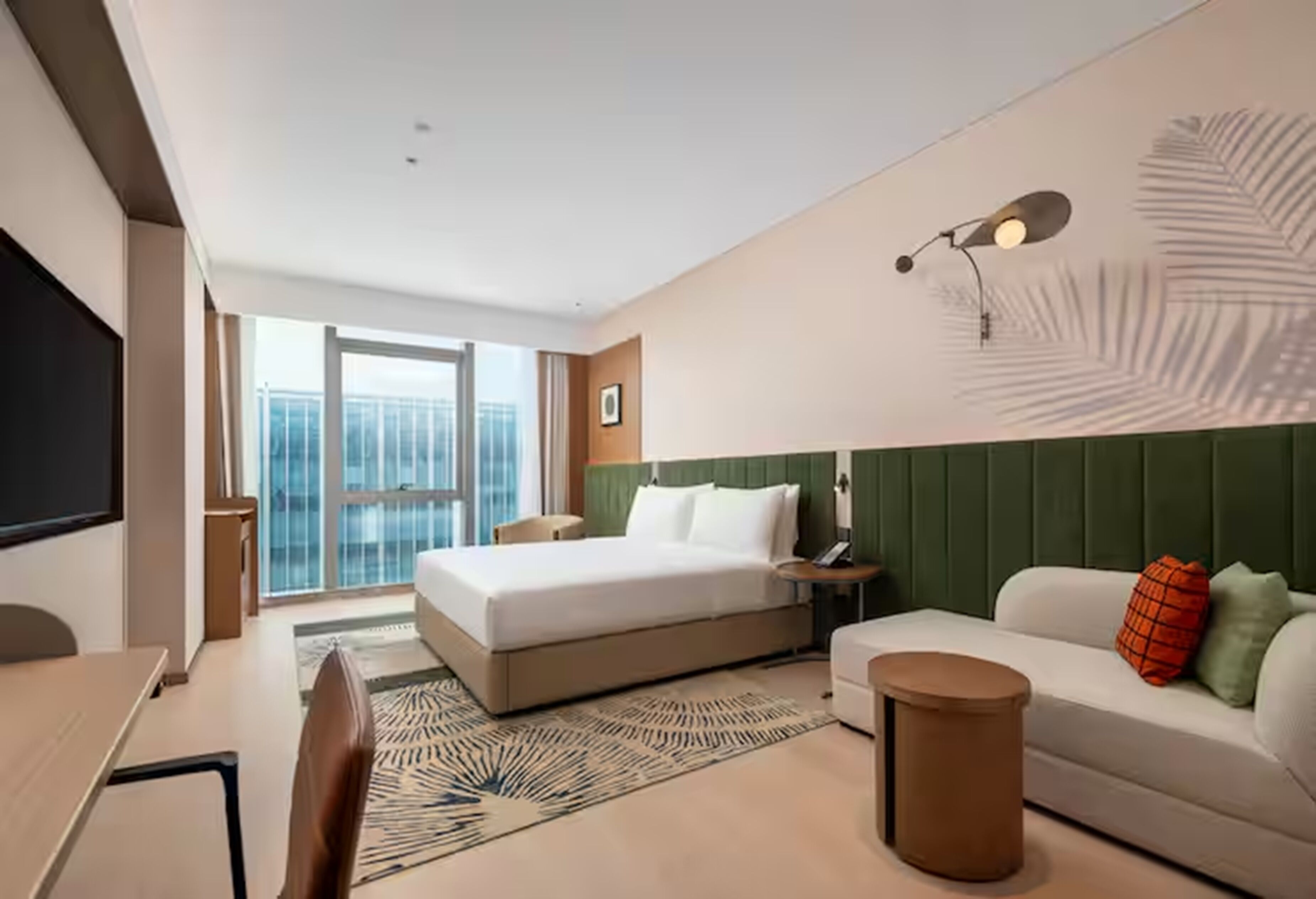 A modern hotel room with a large bed, a desk, a sofa, a round coffee table, a TV, and a balcony with a view.