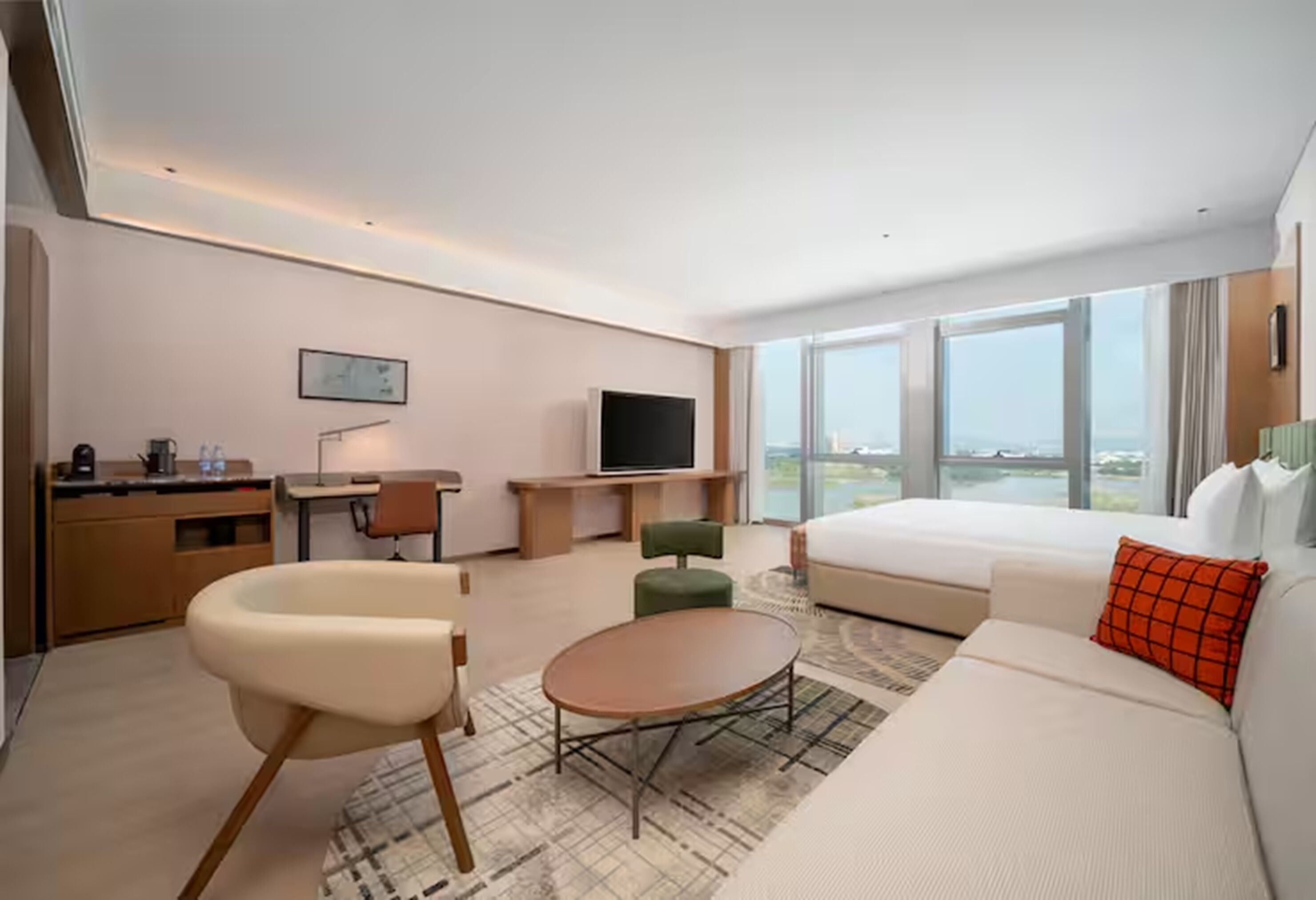 A modern hotel room with a large bed, a sofa, a dining table, a TV, and a view of the city.