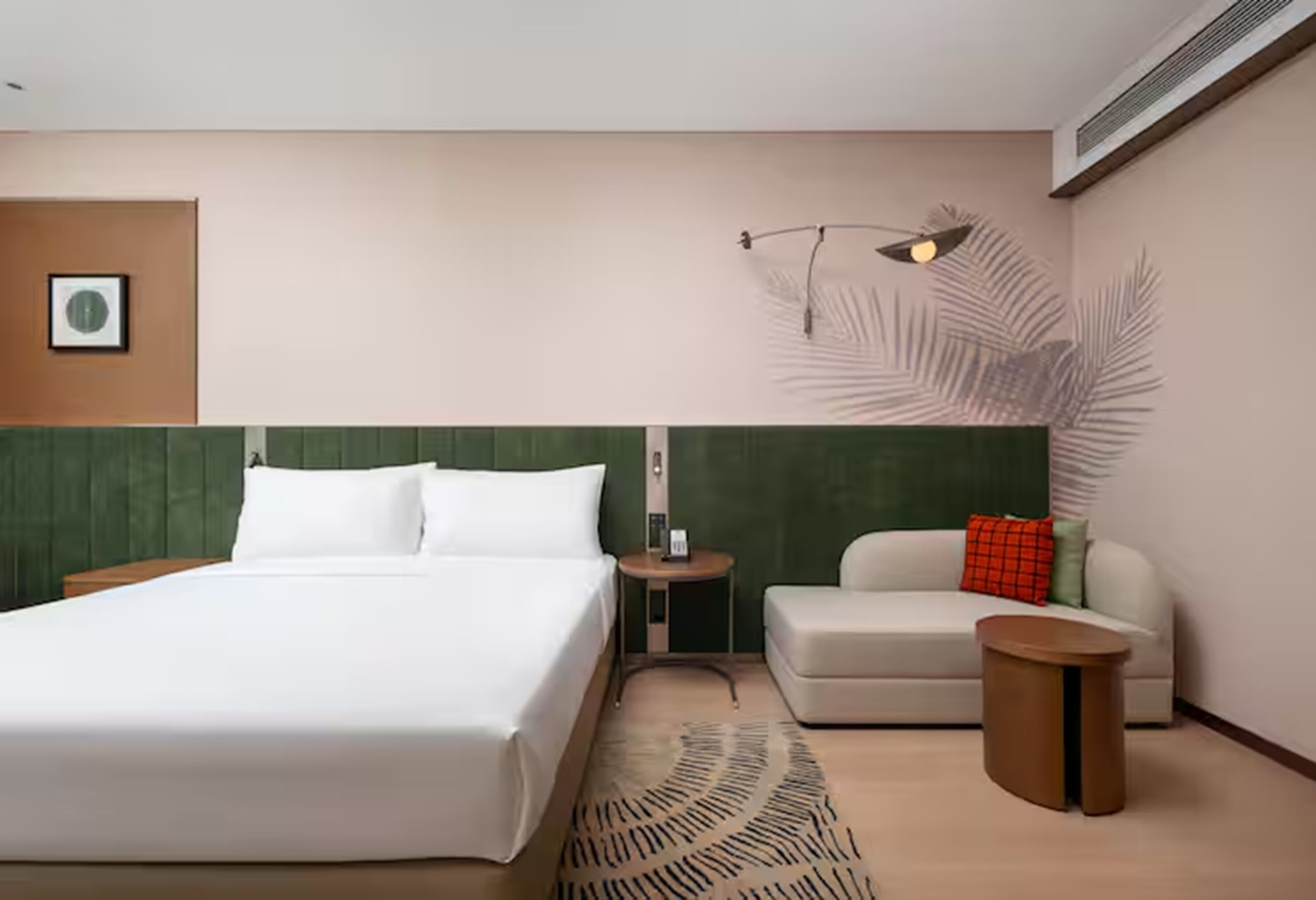 A modern hotel room with a large bed, a sofa, a small side table, and a wall-mounted lamp.