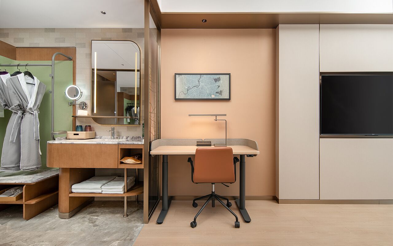 A modern hotel room with a wooden vanity, a desk with a chair, a large mirror, a flat-screen TV, and a framed map on the wall.
