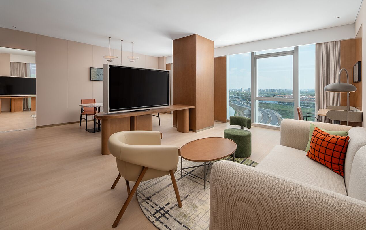 Habitación de hotel moderna con una televisión de pantalla plana grande, un sofá, una pequeña mesa redonda y vistas a un paisaje urbano a través de amplios ventanales.