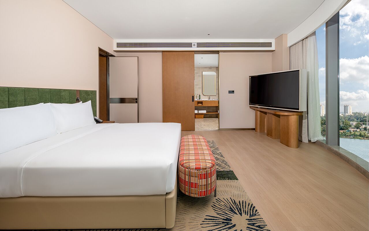 Habitación de hotel con una cama grande, televisión, una otomana pequeña y vistas a la ciudad.