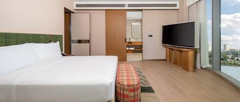 Habitación de hotel con una cama grande, televisión, una otomana pequeña y vistas a la ciudad.