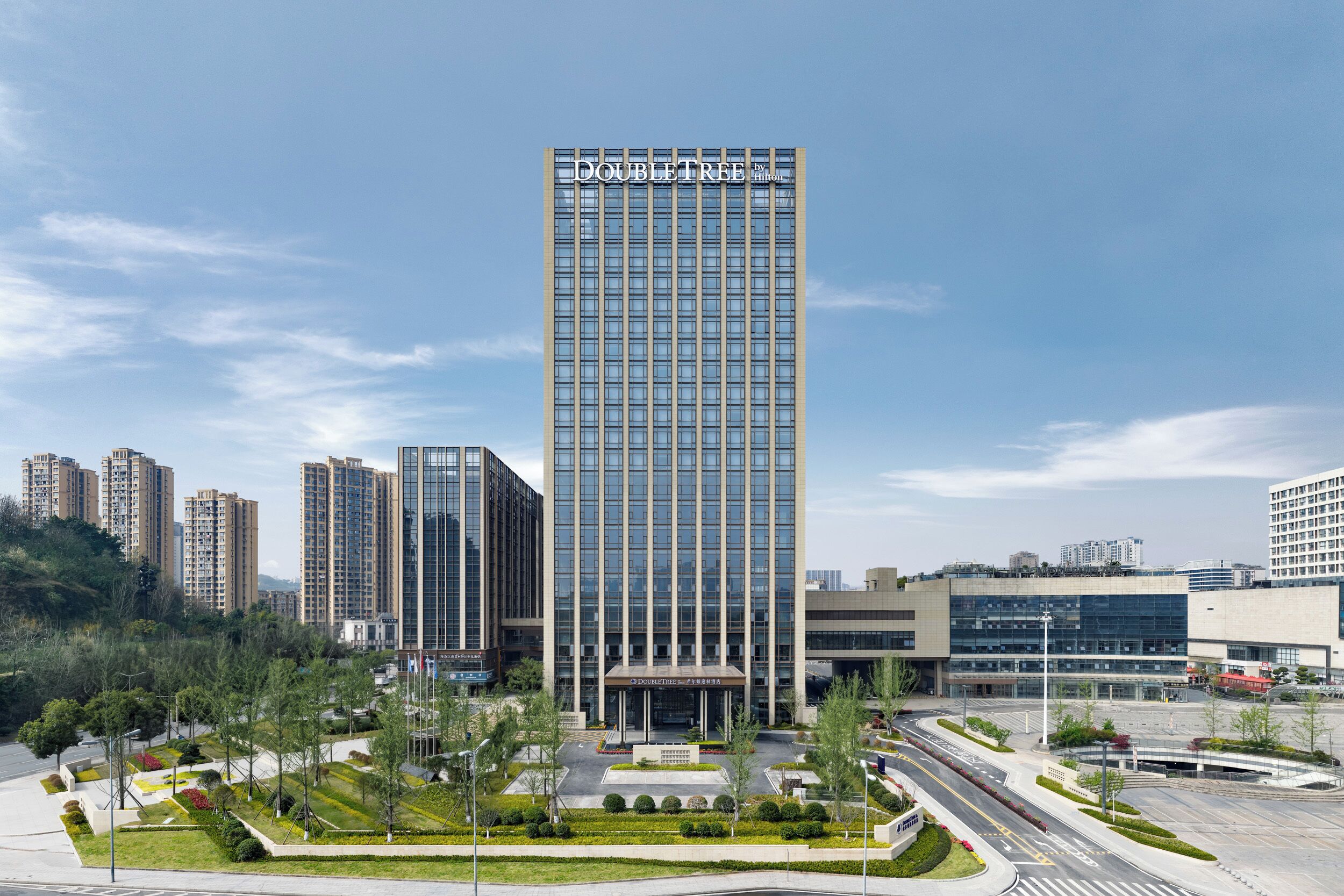 A modern high-rise building with a glass facade, surrounded by landscaped areas and other buildings.