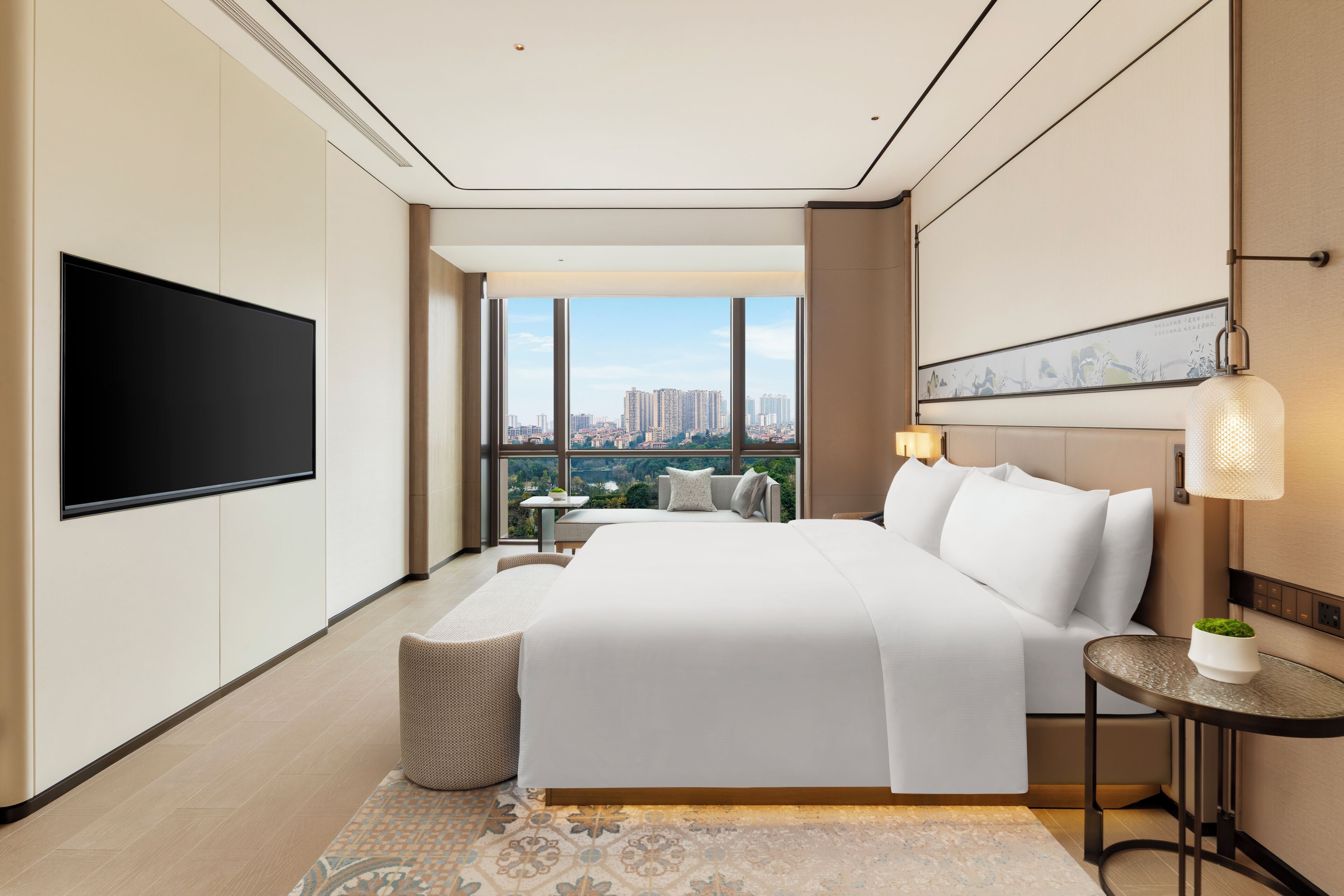 Una habitación de hotel moderna con una cama grande, televisión de pantalla plana, vista a la ciudad, una mesita y una lámpara.