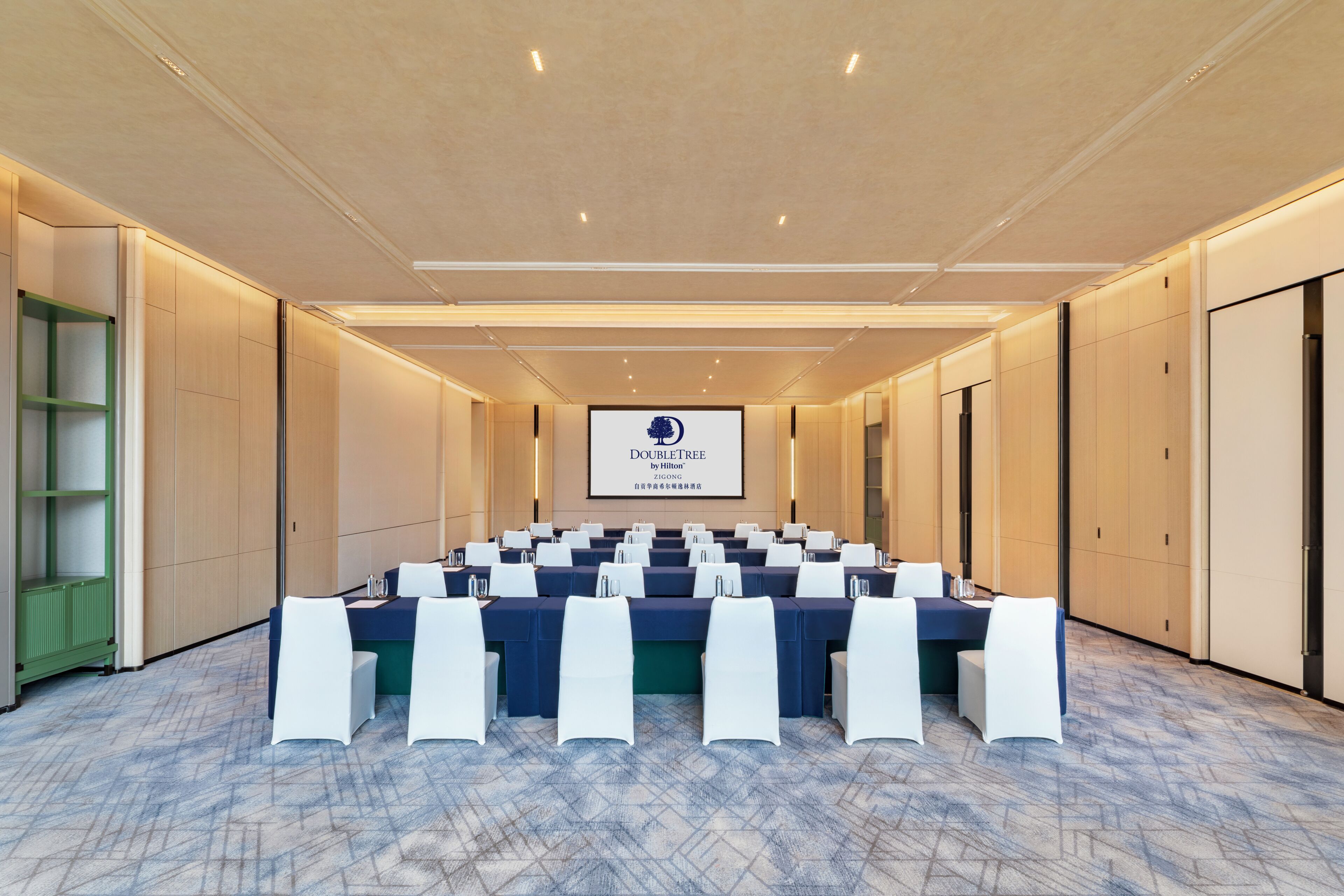 Ein Konferenzraum mit einem langen Tisch, weißen Stühlen und einem Bildschirm, der 'DOUBLETREE by Hilton' anzeigt.