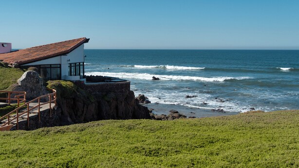 A coastal house with a stone foundation, a red-tiled roof, and a wooden staircase leading to a balcony overlooking the ocean. - La Paloma Rosarito (Playas de Rosarito)